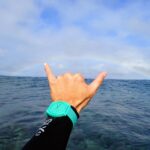 海の上で親指と小指を立ててシャカサイン（アロハサイン）を出す手。背景に虹がかかる